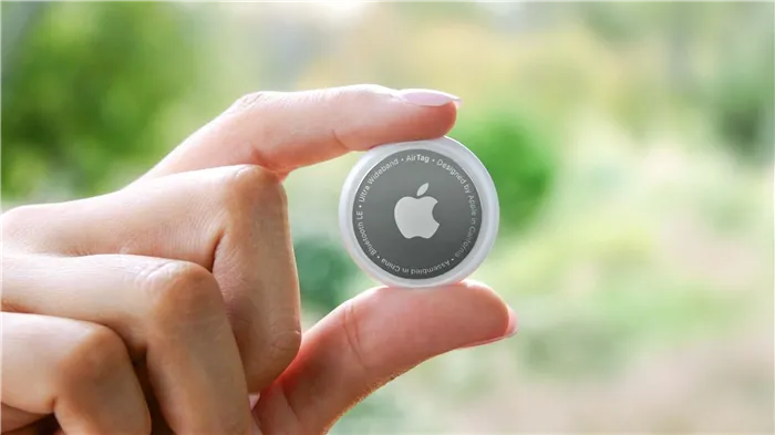 Apple AirTag, зажатый между пальцами пользователя на размытом зеленом фоне.
